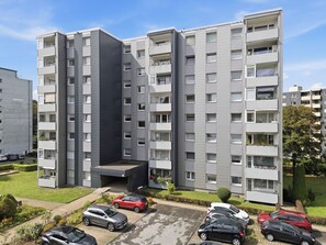 A multi-story residential building with balconies, a parking area with several cars, and a well-maintained lawn.
