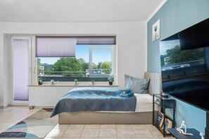 A bedroom with a bed, a TV, a window with blinds, and a view of greenery outside.