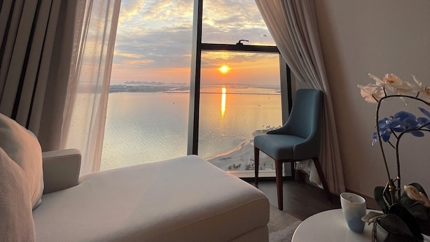 A modern hotel room with a large window offering a sunset view over a body of water. There is a sofa, a chair, and a small table with a vase of flowers. - Sea view Àlacarte Ha Long Bay Apartment (Bai Chay)