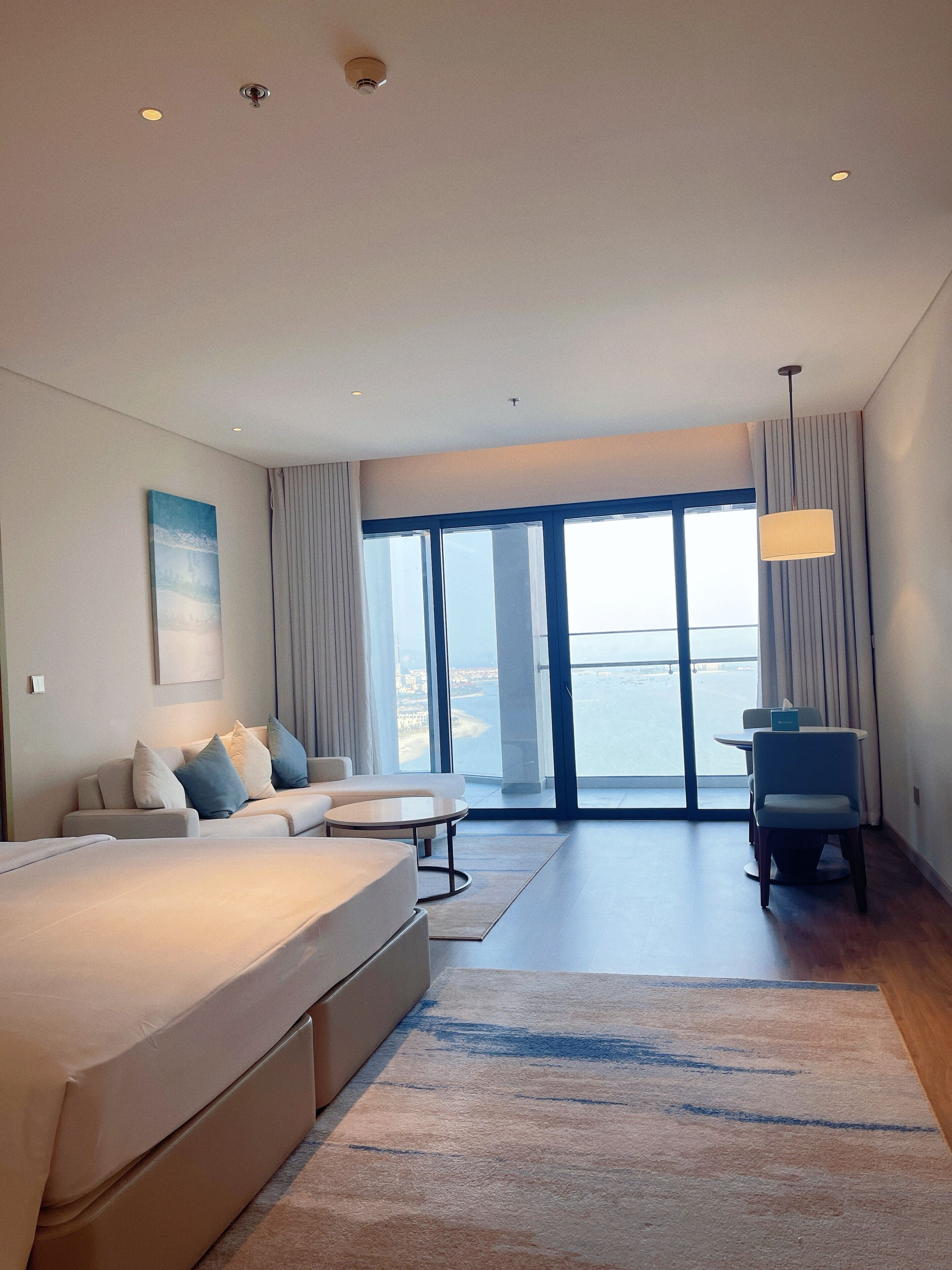 A modern hotel room with a large bed, a seating area with a table, and a view of the sea through large windows.