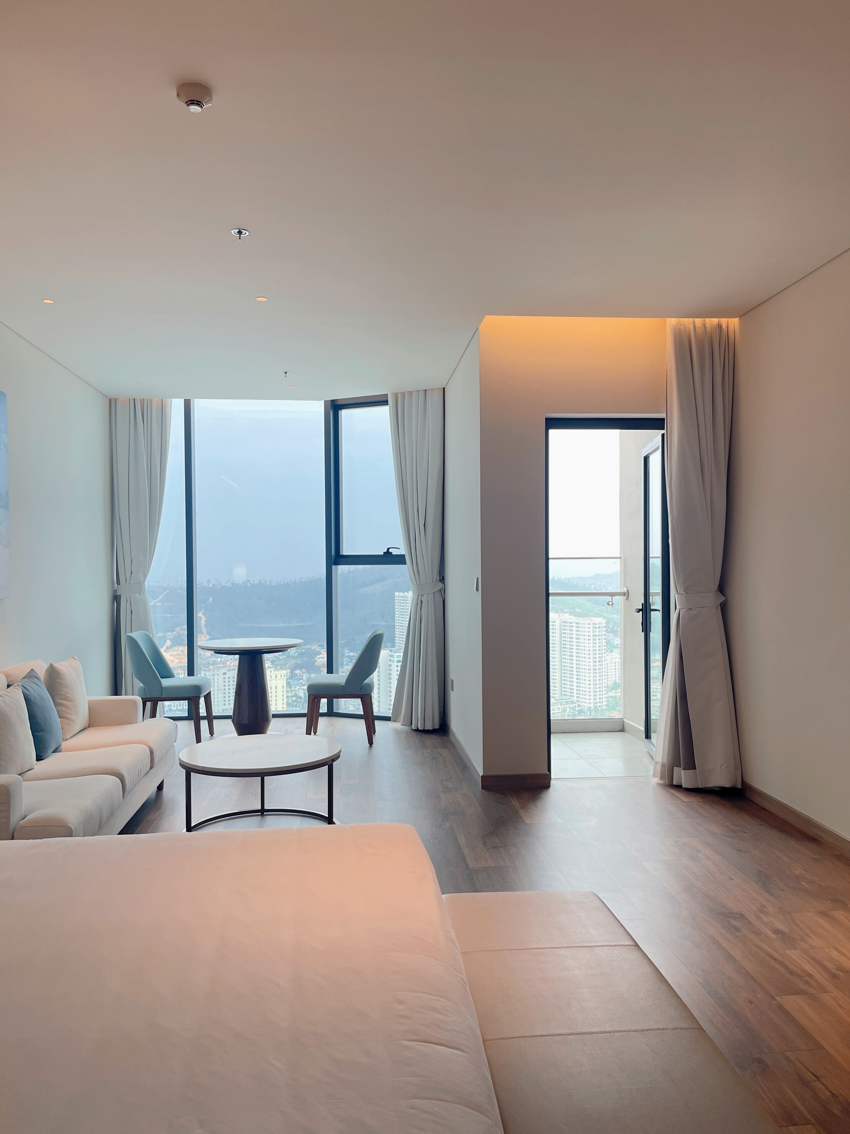 A modern hotel room with a large window, a sofa, two chairs, a small table, and a view of the city.