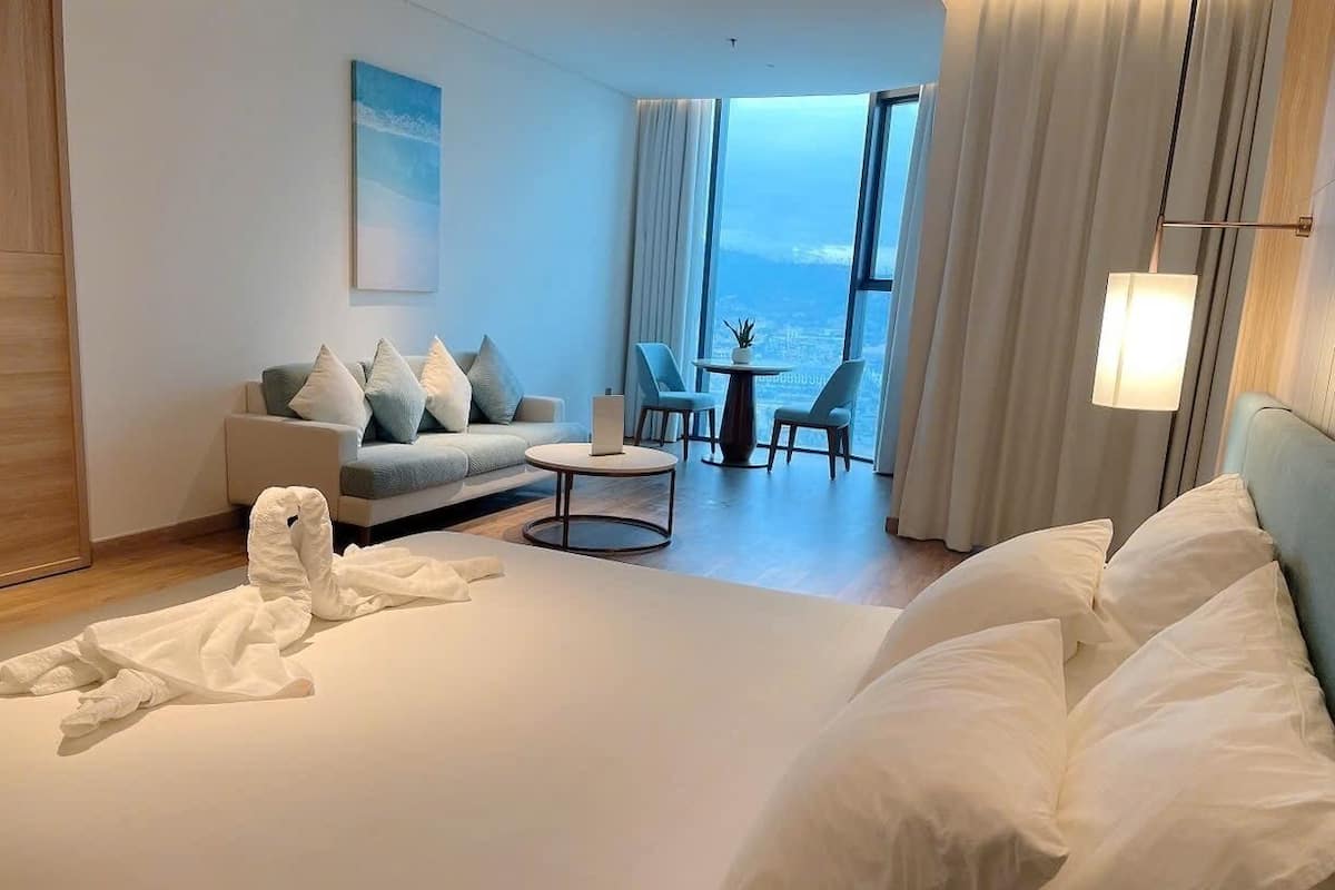 Habitación de hotel con cama, sofá, una mesita y vistas a la ciudad a través de amplios ventanales.