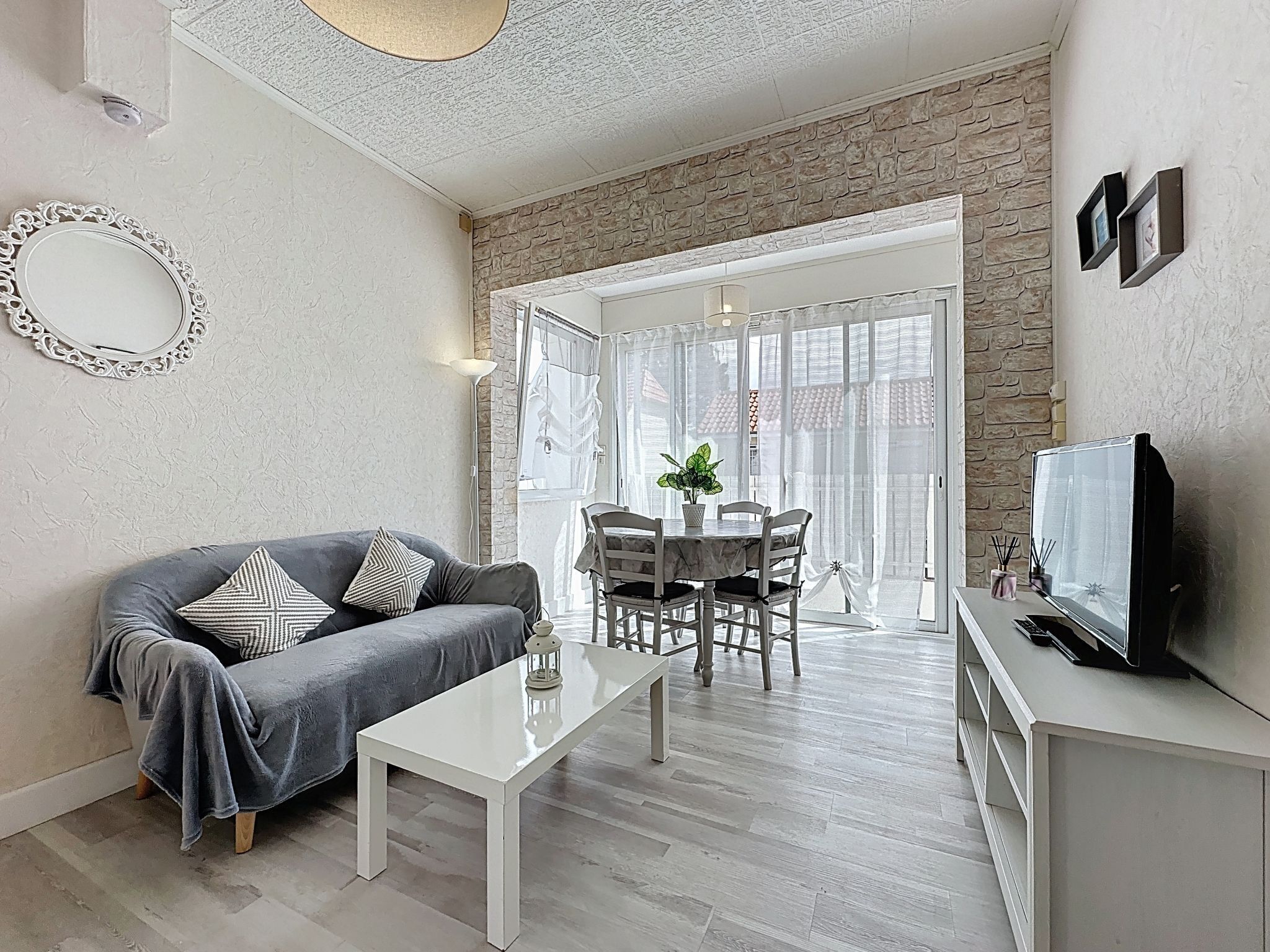 Una sala moderna con un sofá gris, una mesa de centro blanca y un comedor con mesa y sillas. Un televisor está sobre un mueble a la derecha.