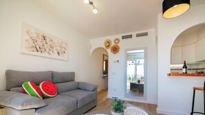 Una sala de estar moderna con un sofá gris, una mesa de centro con felpudos decorativos, un cuadro y vistas al área de la cocina.