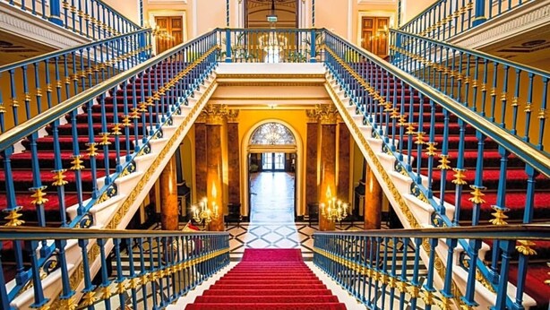 A grand staircase with red carpet and blue balustrades, leading to a grand hall with chandeliers.