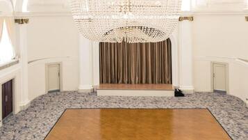 A spacious room with a large wooden floor, a grand chandelier, and a stage with curtains.