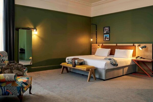 Habitación de hotel con una cama, un banco, una silla, una mesita de noche y un espejo.