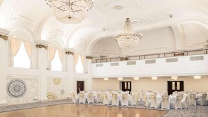 A large hall with rows of tables and chairs, high ceilings with chandeliers, and a polished floor.