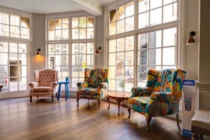 Una habitación amplia con ventanas grandes, un sillón clásico, sillones coloridos y una pequeña mesa redonda.