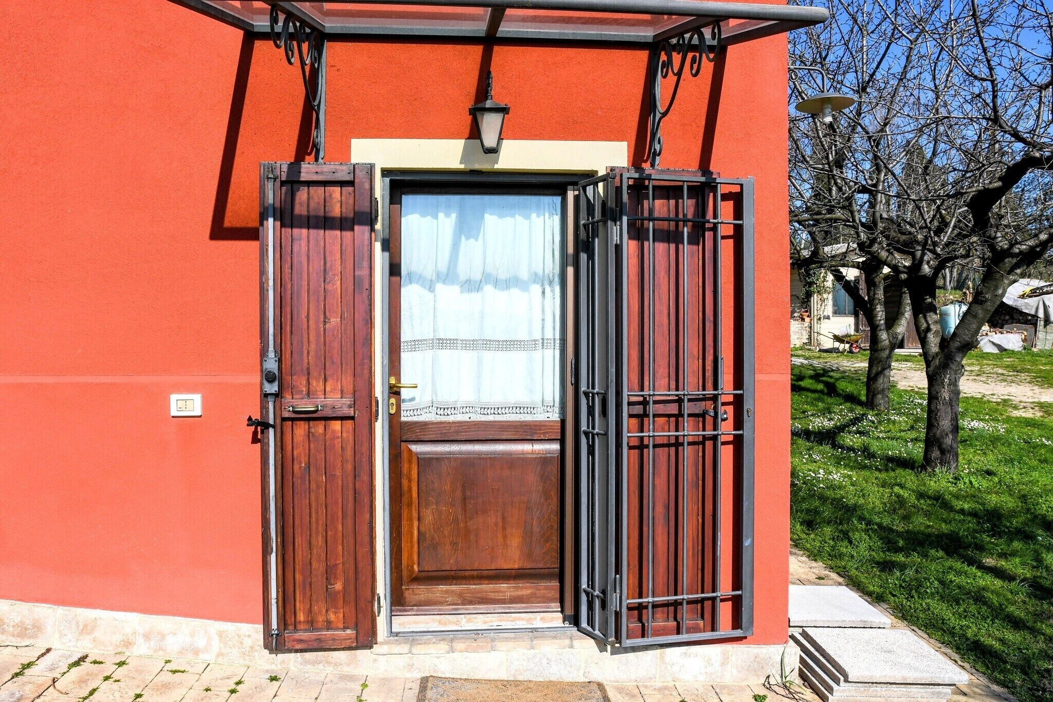 A red building with a wooden door and a metal gate, a light fixture above the door, and a tree in the background.