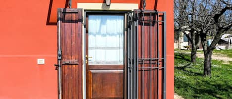A red building with a wooden door and a metal gate, a light fixture above the door, and a tree in the background.