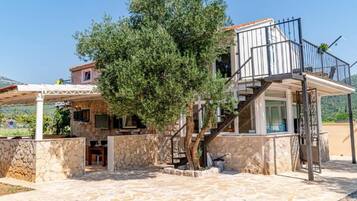 A stone-walled building with a staircase leading to a balcony, surrounded by a garden and a view of the countryside.