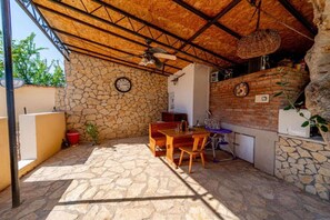 Un patio techado al aire libre con suelo de piedra, una mesa y sillas de madera, un muro de ladrillo y un reloj.