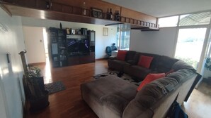 Uma sala de estar com sofá, televisão e mesa de centro. Há uma prateleira de madeira acima do sofá e uma janela com vista externa.