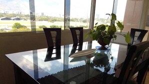 Mesa de jantar com cadeiras e um vaso de planta, grandes janelas com vista para um parque e edifícios.