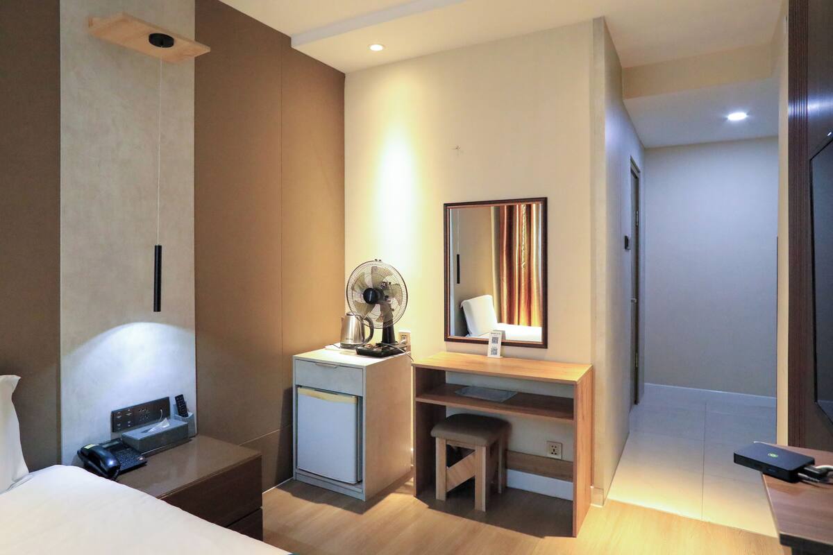 A hotel room with a bed, a desk with a fan, a small cabinet, a mirror, and a television.