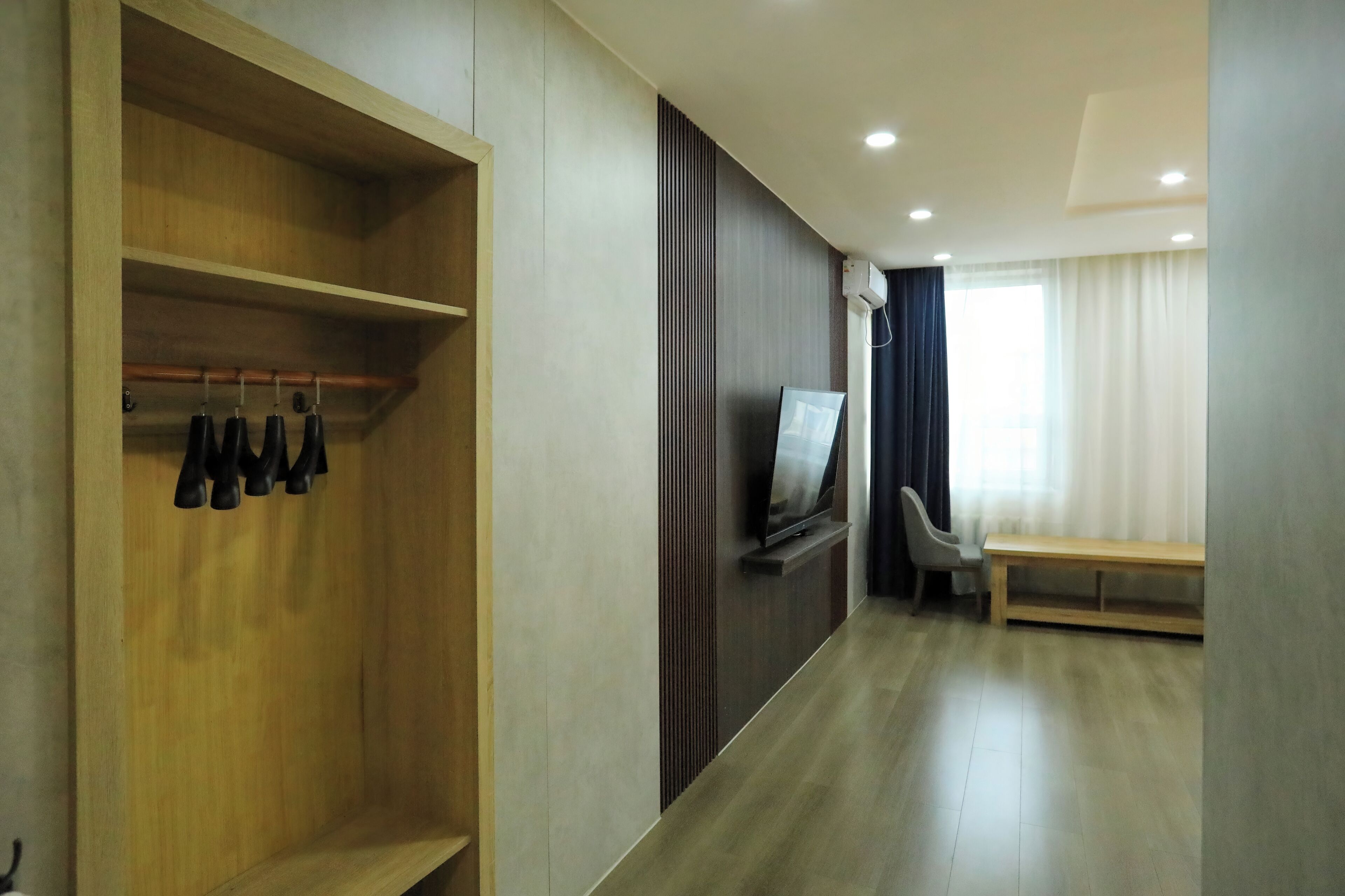 Habitación de hotel con armario de madera, televisor montado en la pared, una banca y ventana con cortinas.