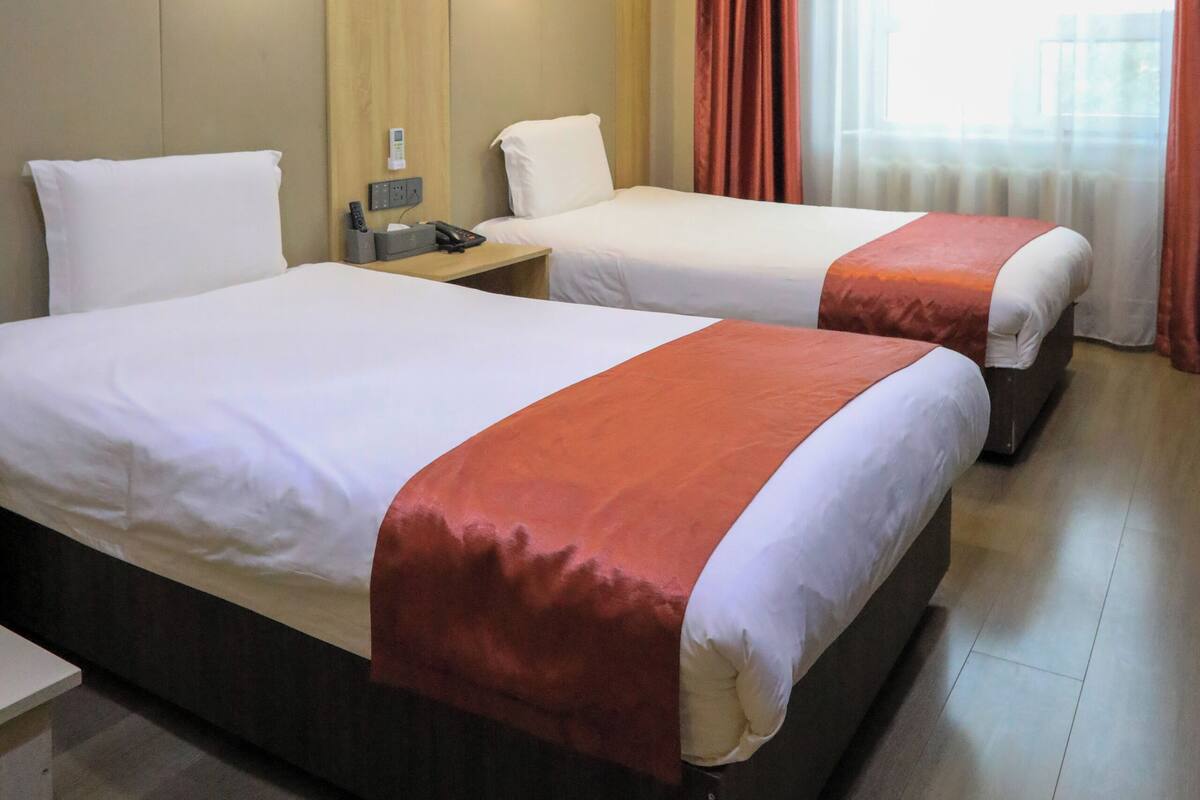 A hotel room with two beds, a bedside table with a phone and charger, a window with curtains, and a wooden wall panel.