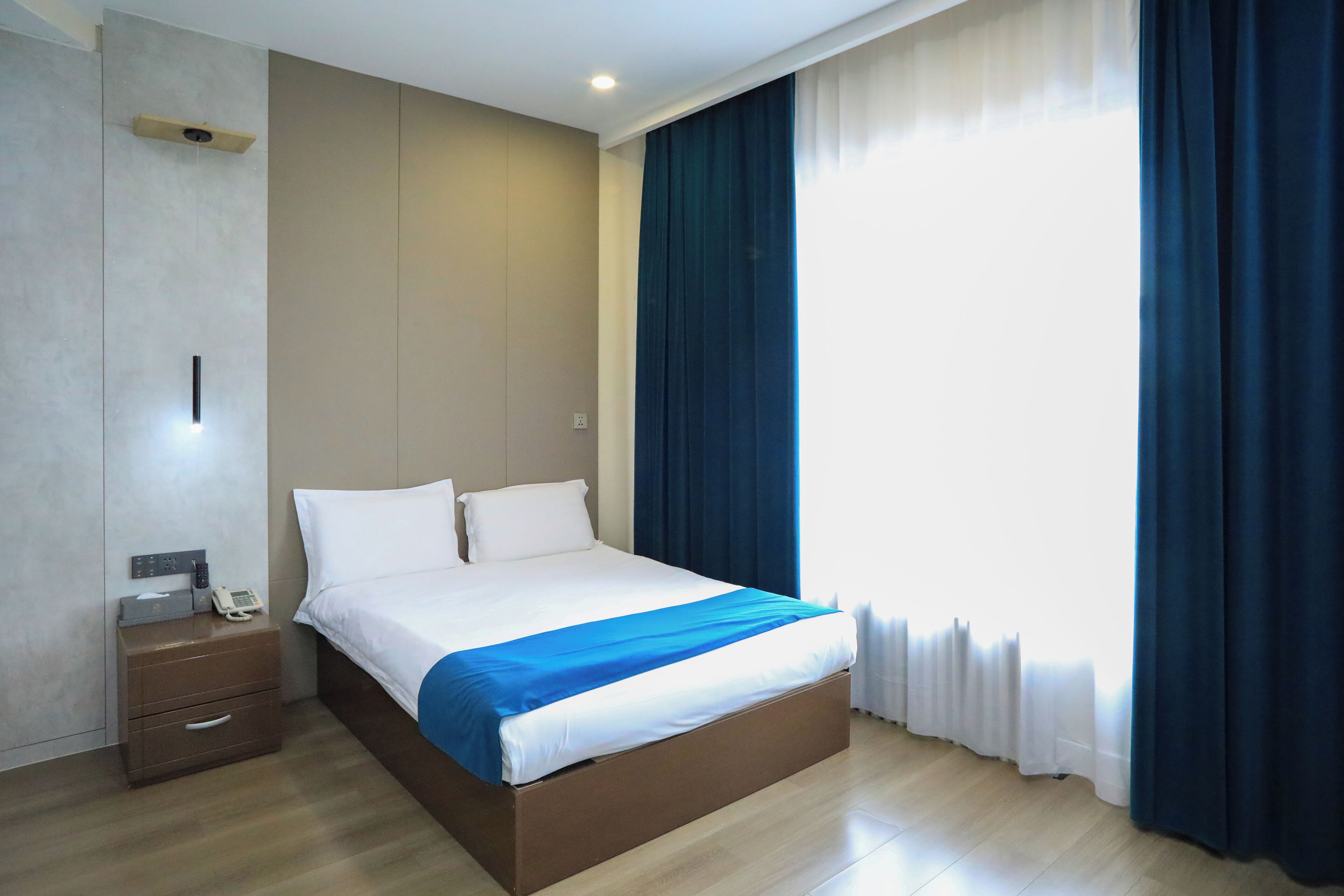 A neatly made bed with a blue accent, a bedside table with a phone, and a wall-mounted light fixture.