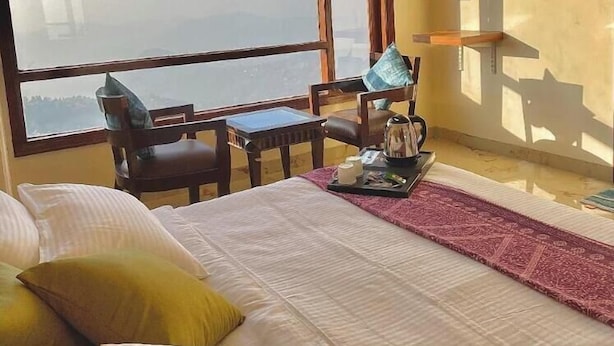A bedroom with a large window, a bed with pillows, a small table with a kettle, and a chair. - Hotel The Waypoint Inn (Shimla)