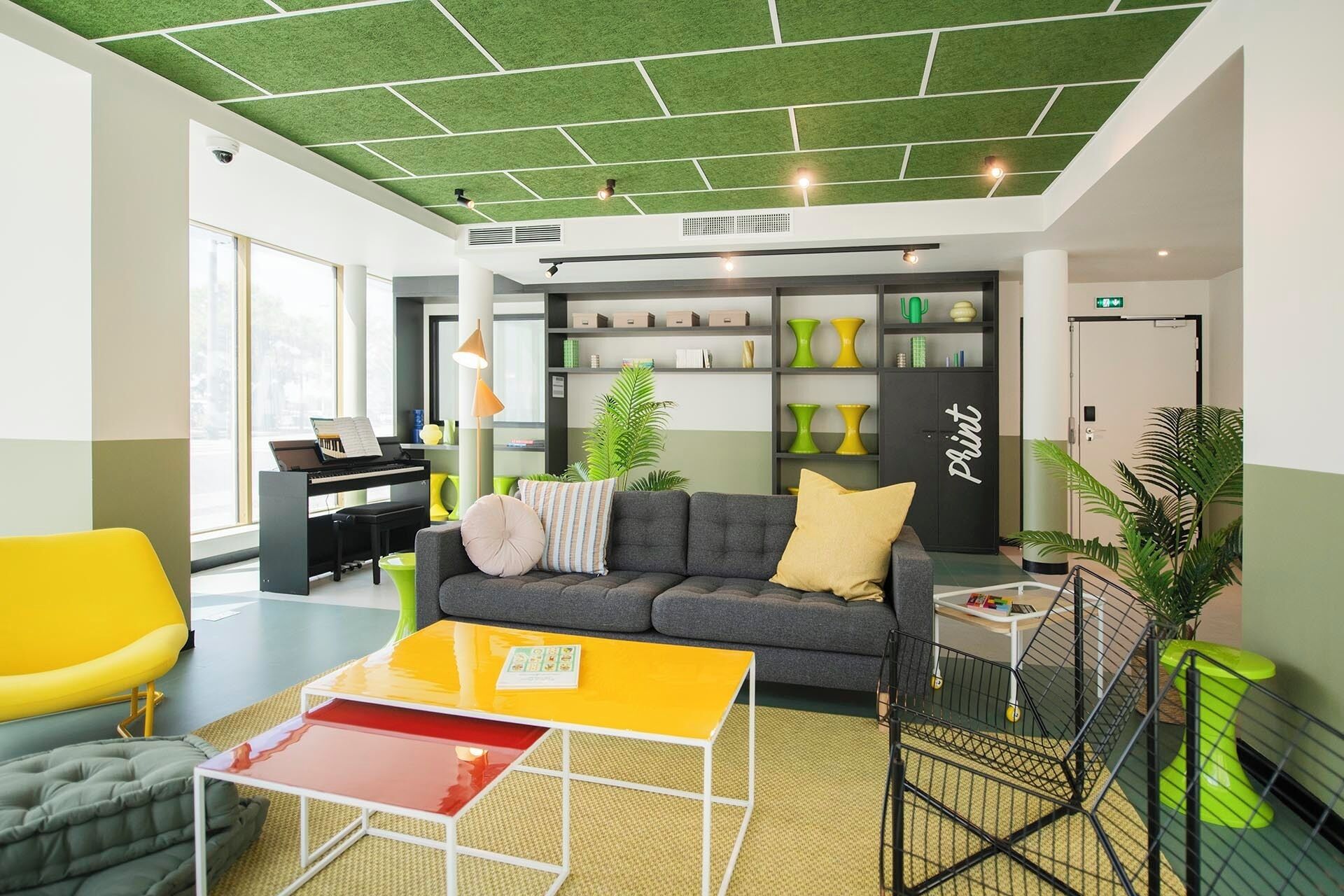 Un vestíbulo moderno con techo de baldosas verdes, un piano, un sofá con cojines, una mesa amarilla y una estantería con jarrones decorativos.