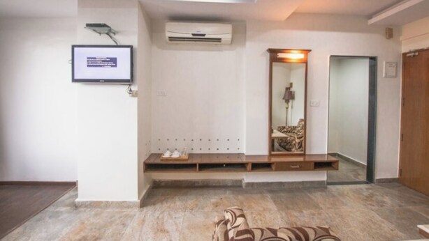 A modern interior with a flat-screen TV mounted on the wall, a built-in bench with storage, a mirror, and a door to the right. - Hotel Sarayu Hassan (Hassan)