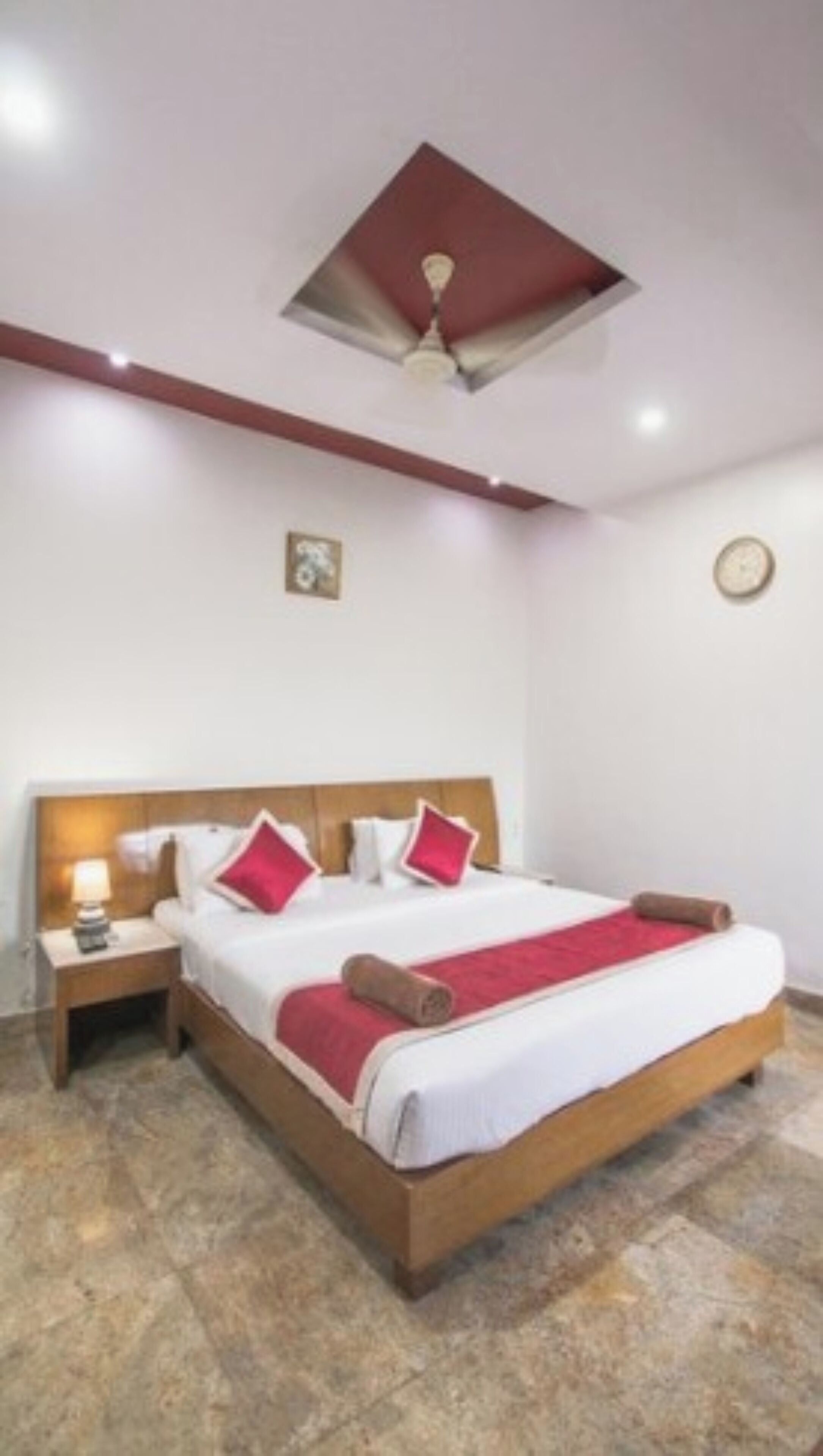 A bedroom with a wooden bed, white bedding, red pillows, a red throw blanket, a nightstand with a lamp, a ceiling fan, and a clock on the wall.