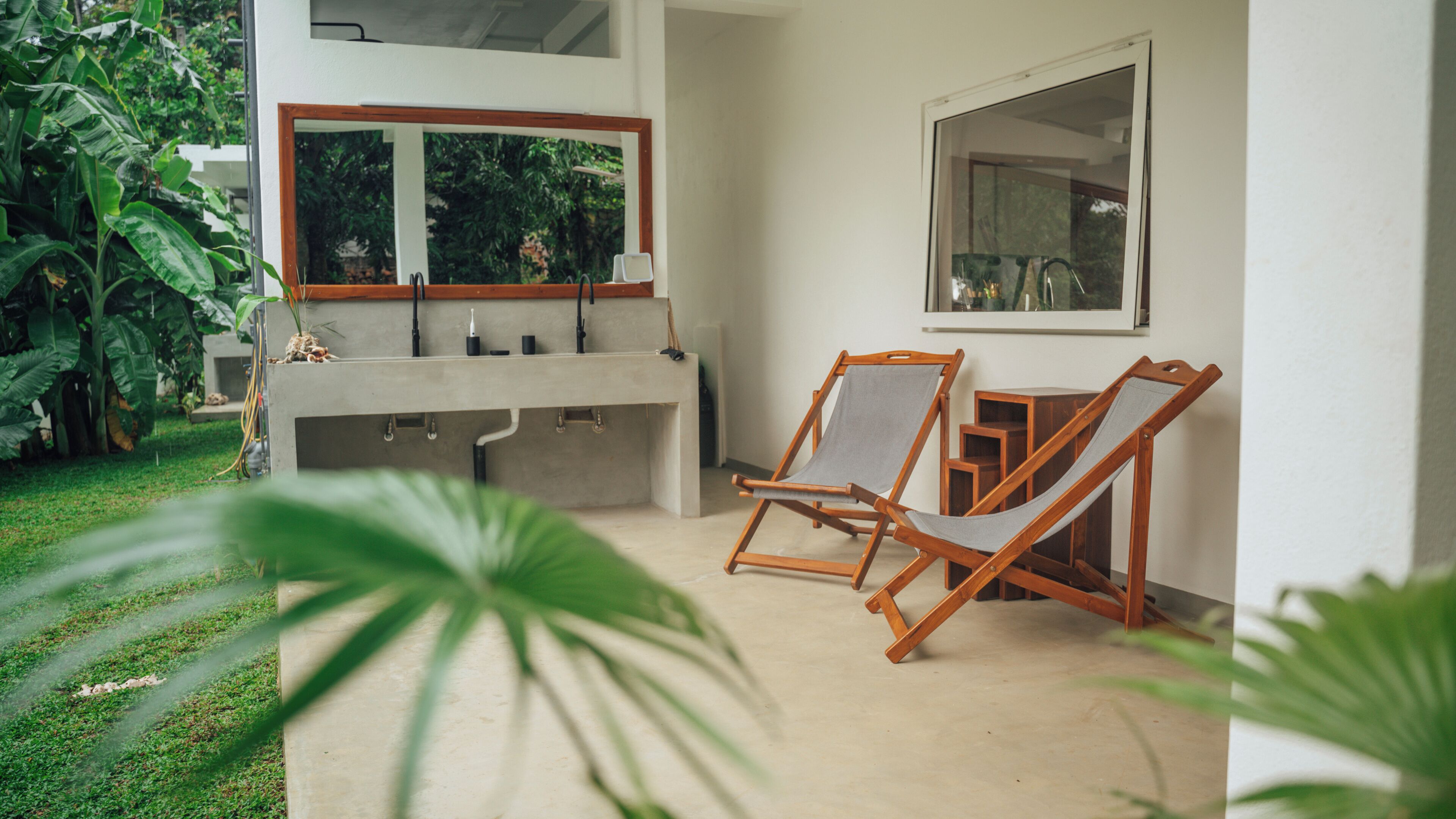 A modern outdoor kitchen area with a concrete countertop, two wooden chairs, and a large window overlooking greenery.