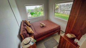 A small, neatly arranged bedroom with a single bed, two windows, a wooden nightstand, a hat, and a small table with a book and a decorative item.