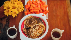 A plate of pancakes with two cups of coffee, a bowl of cubed fruit, and a vase of yellow flowers on a wooden table.