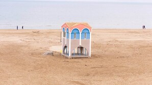 Una petita cabana de platja colorida amb teulada de rajoles i finestres arquejades, situada a una platja sorrenca amb un mar tranquil al fons.