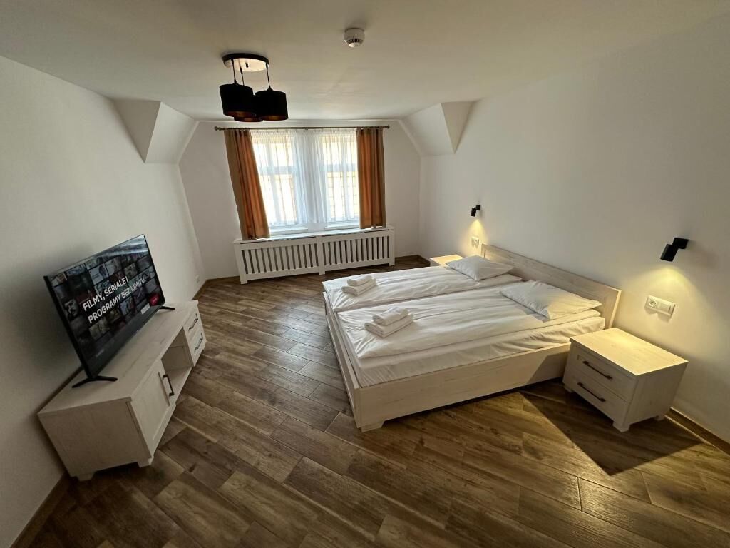 A bedroom with a large bed, two bedside tables, a TV on a stand, a window with curtains, and a radiator.