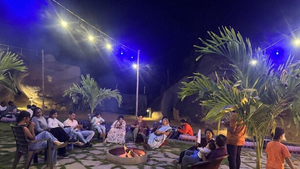 A group of people seated around a fire pit in an outdoor setting with stone flooring and palm trees. - HILL STONE VIEW HAMPI (Koppal)