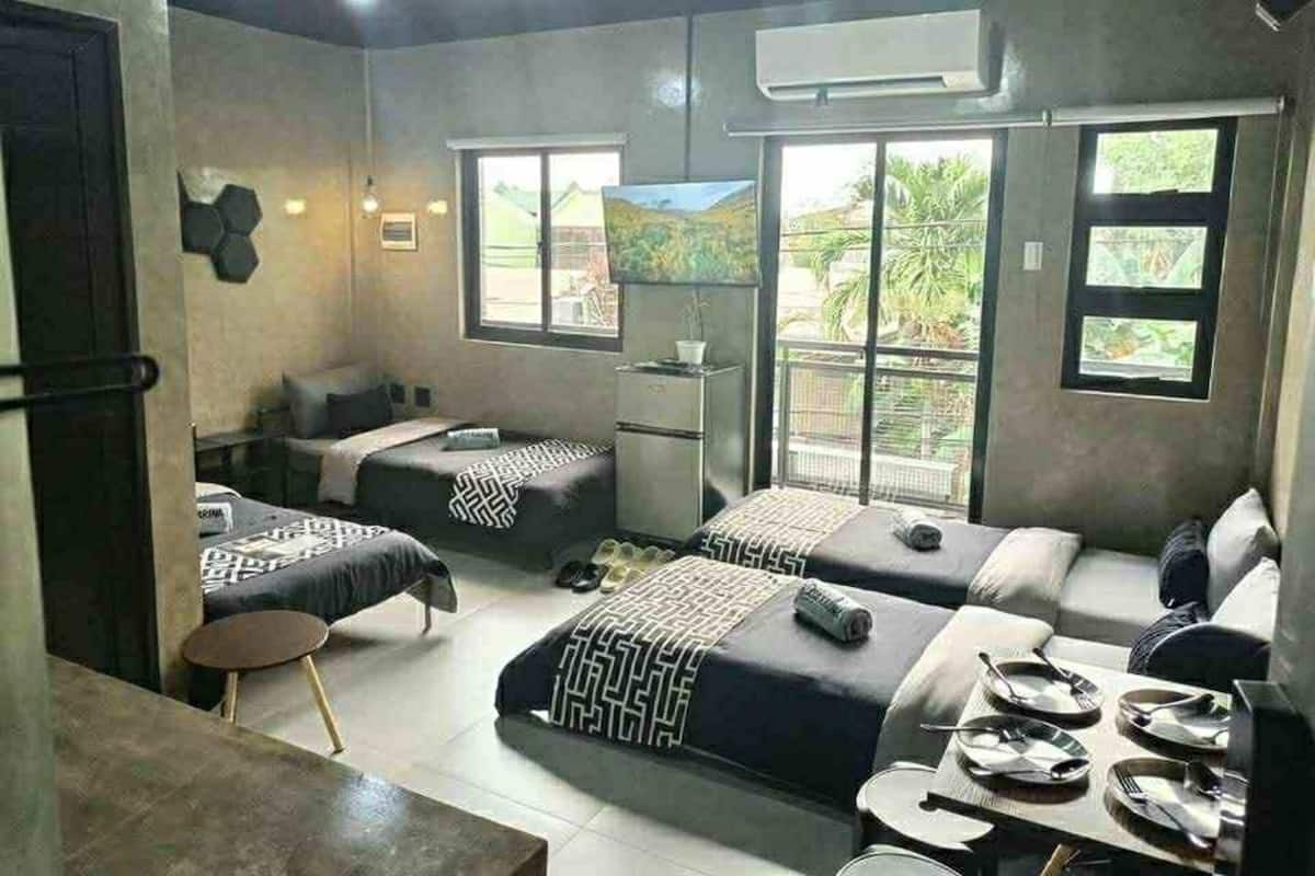 A room with two beds, a small table, and a TV. There are windows with a view of greenery outside.