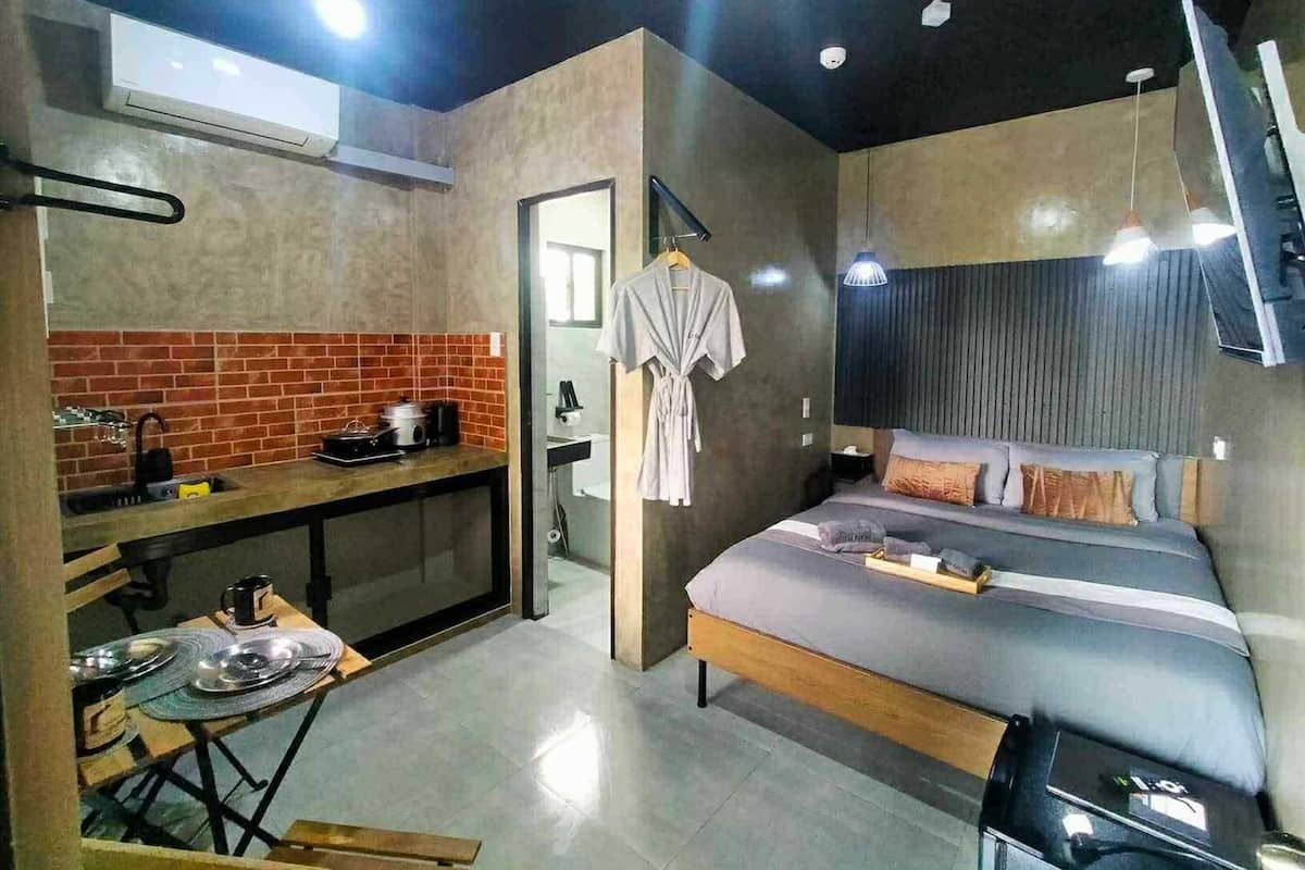A compact hotel room with a kitchenette, a bed with pillows, a TV, and a ceiling-mounted air conditioner.
