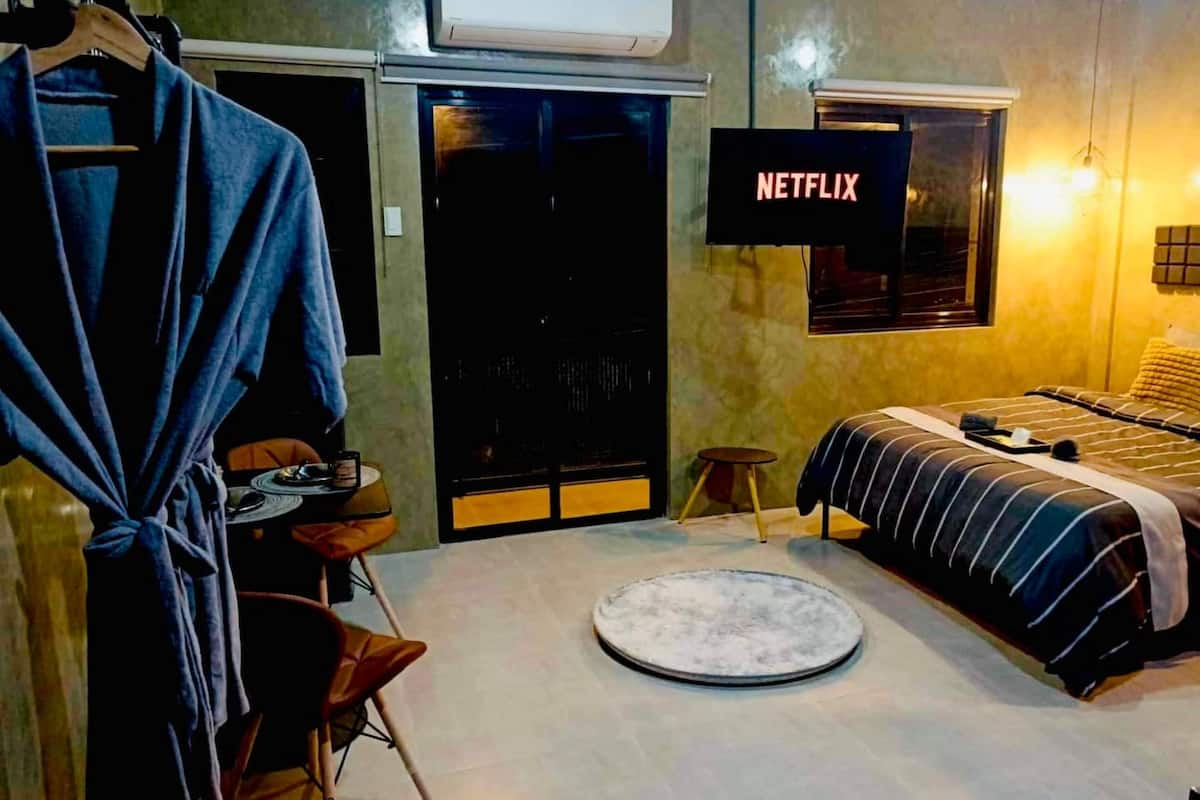 A bedroom with a bed, a round rug, a chair, a small stool, a robe hanging, and a TV displaying 'NETFLIX'.