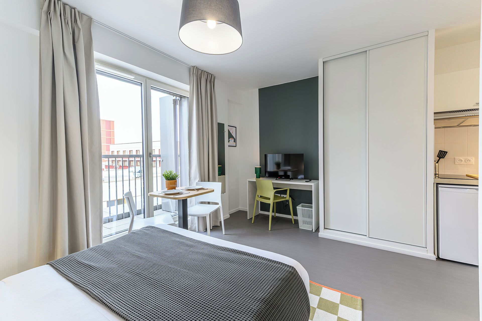 Una habitación de hotel moderna con cama, escritorio con computadora, kitchenette pequeña y balcón con cortinas.