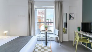 A modern hotel room with a bed, a desk, a chair, a TV, and a balcony with a view of buildings. - Student Factory Paris - Universeine  (Saint-Denis)