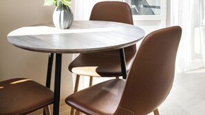Una mesa redonda de comedor con cuatro sillas marrones, un florero con flores rosas, y vista a un crucero a través de una ventana con cortinas blancas.