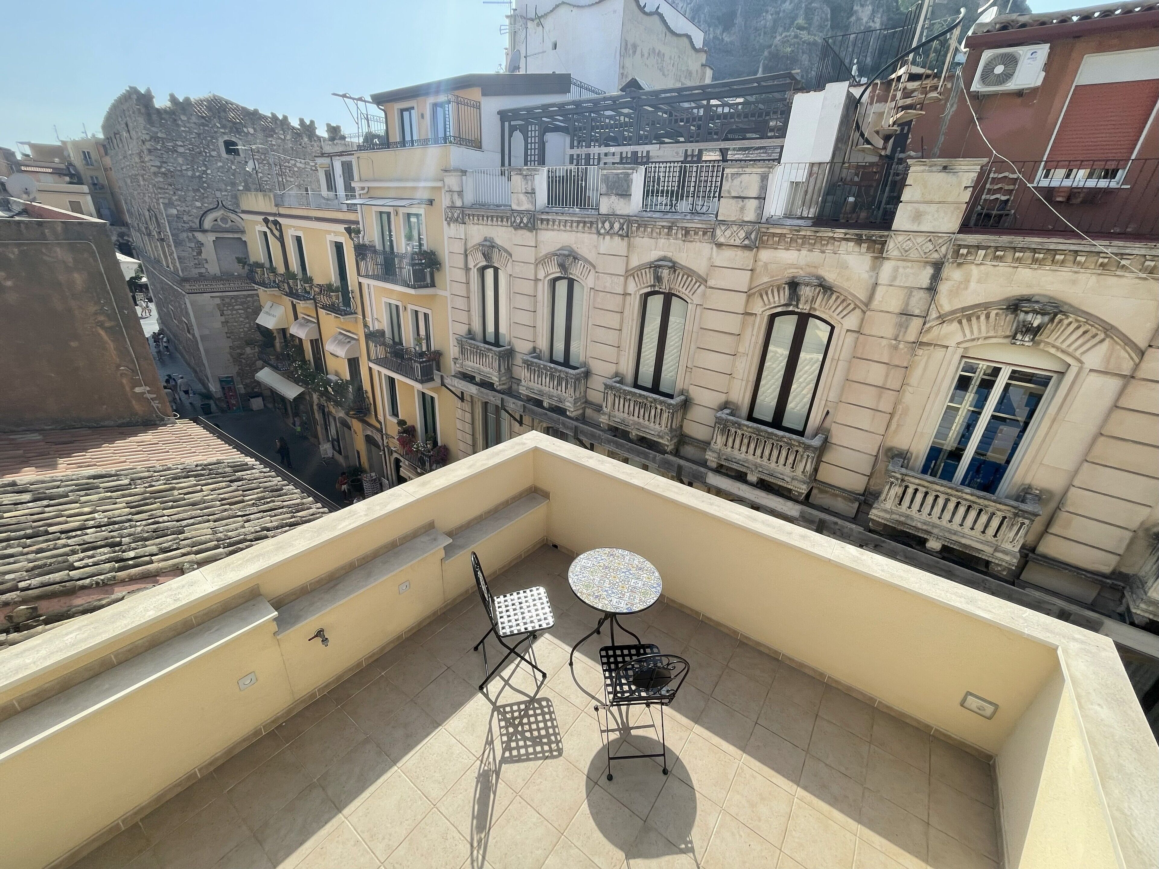 A balcony with two metal chairs and a small round table, overlooking a street with historic buildings and a stone wall.