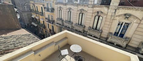A balcony with two metal chairs and a small round table, overlooking a street with historic buildings and a stone wall.