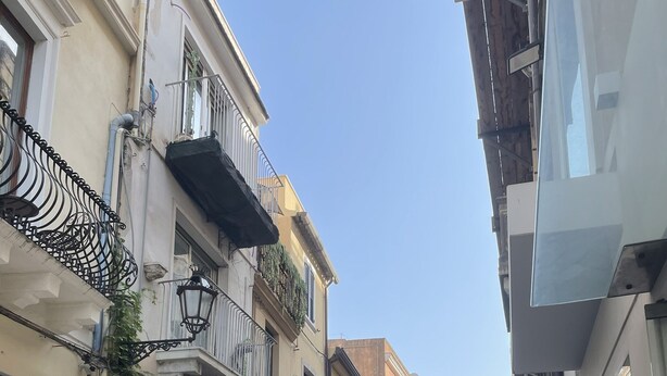 Une rue étroite bordée de bâtiments des deux côtés, avec des tables et des chaises installées à l’extérieur d’un café, et des piétons qui se promènent.
