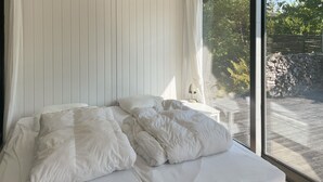 Ein Schlafzimmer mit großem Fenster, weißer Bettwäsche und Blick auf eine Steinmauer und Grünpflanzen.