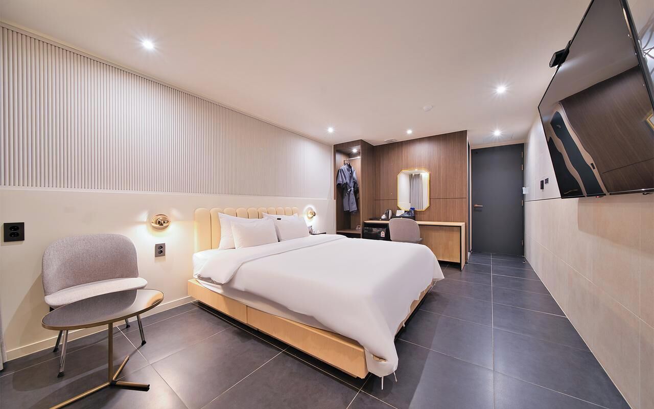 A modern hotel room with a large bed, a bedside table, a chair, a vanity with a mirror, and a wardrobe.