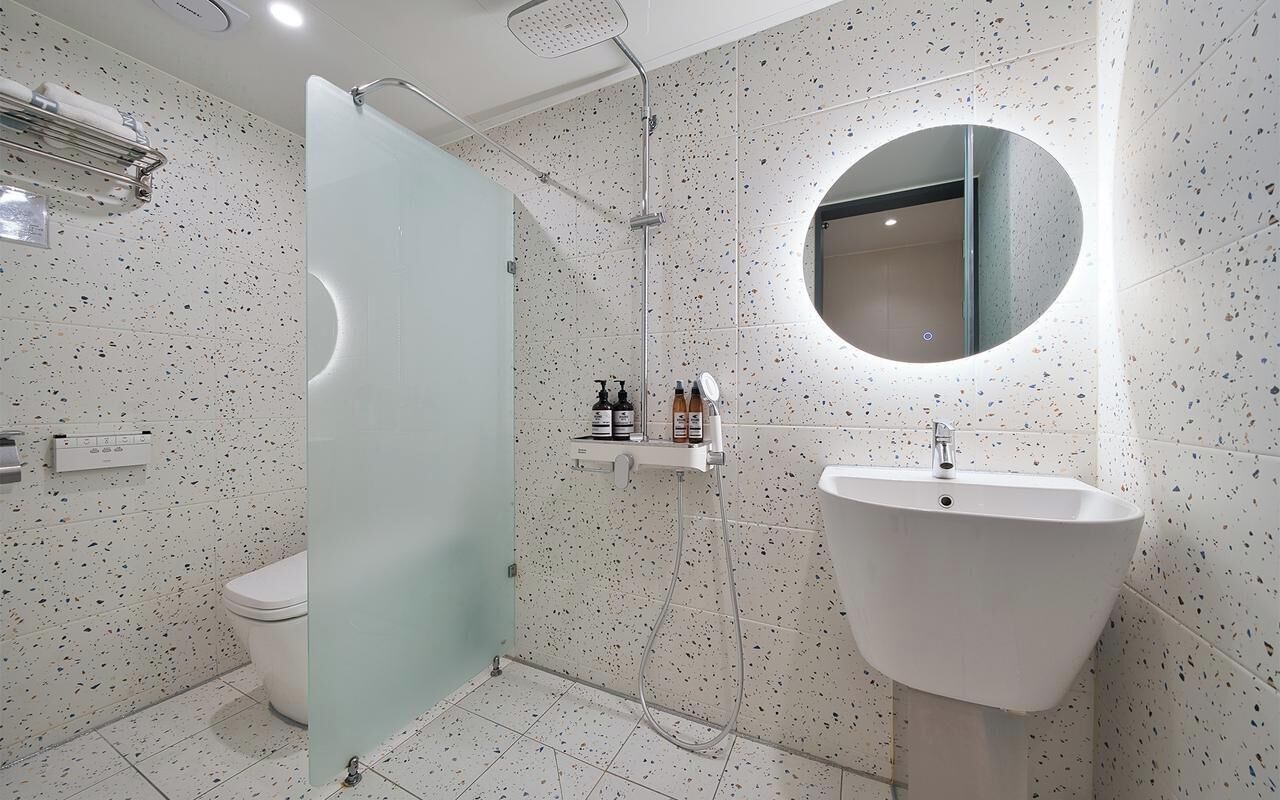 A modern bathroom with a freestanding toilet, a glass shower enclosure, a round mirror, and a white sink.