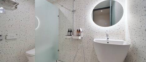 A modern bathroom with a freestanding toilet, a glass shower enclosure, a round mirror, and a white sink.