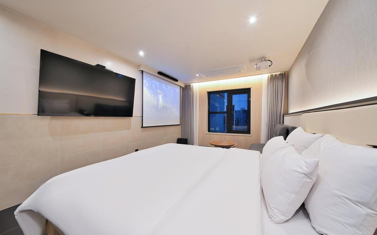Une chambre d’hôtel moderne dotée d’un grand lit, d’un téléviseur à écran plat fixé au mur, d’un écran de projection et d’une fenêtre donnant sur la ville.