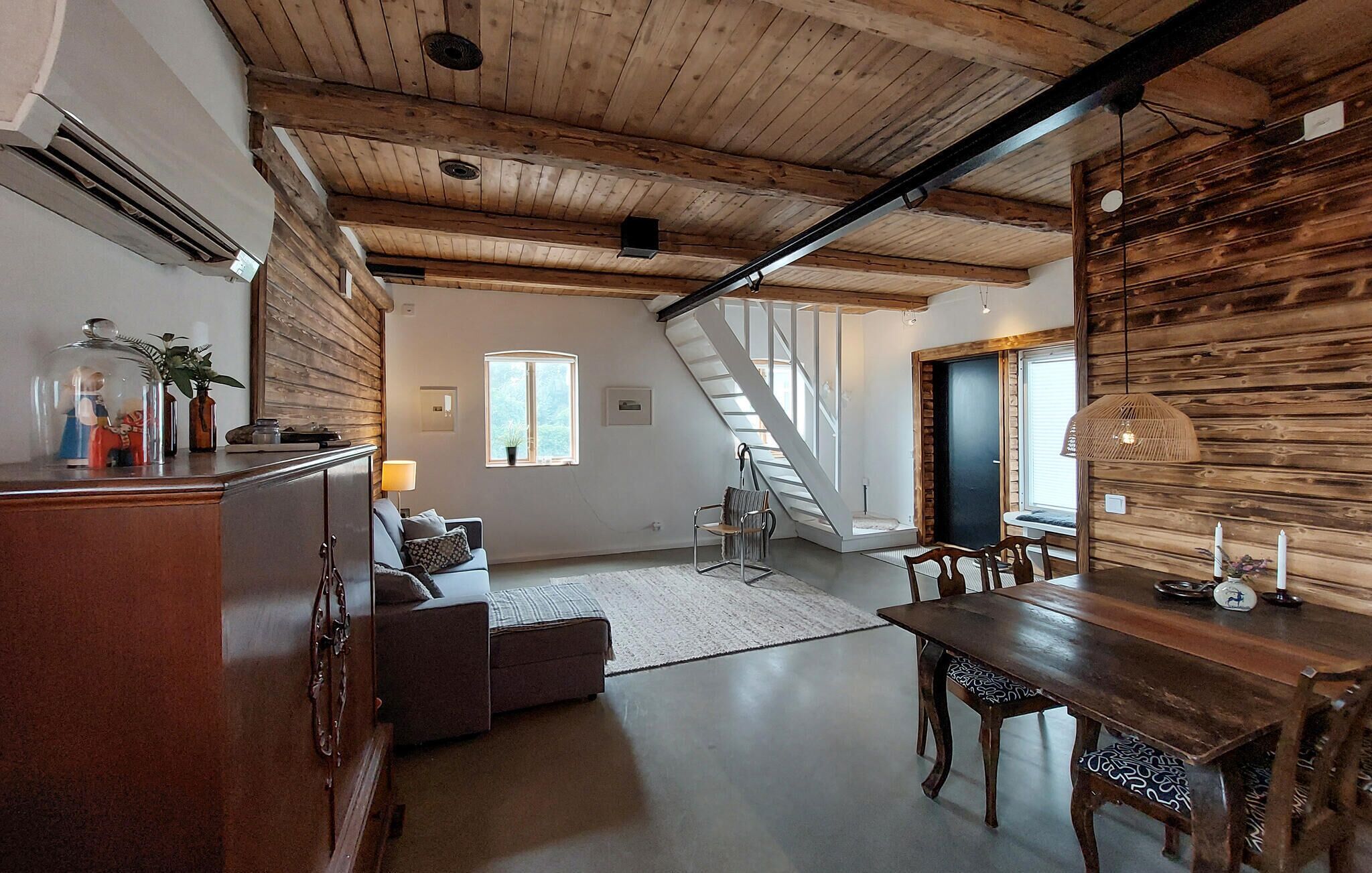 A spacious loft with a high ceiling, exposed wooden beams, a staircase, and a dining area with a wooden table and chairs.