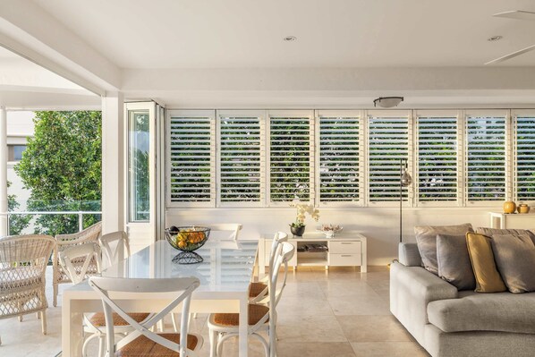 A modern living room with a glass dining table, white chairs, a grey sofa, and a large window with blinds.