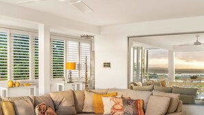 A modern living room with a beige sectional sofa, a glass coffee table, and large windows with white shutters. - Picture Perfect - Panoramic Views, Walk to Beach (Noosa Heads)